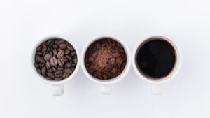 Three cups, one with coffee beans, second with ground coffee and the third with a coffee drink