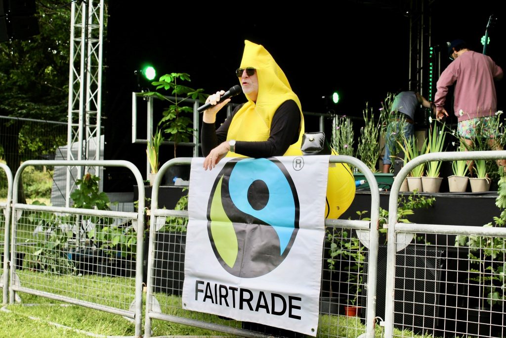 A man in a banana costume wearing sunglasses addresses a crowd from a stage, which is full of plants.